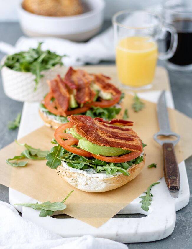blt fried egg bagelwich, Two toasted bagels topped with dairy free cream cheese arugula tomato avocado and crispy bacon sitting on a marble cutting board