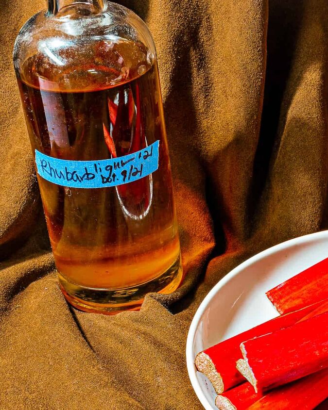 easy homemade rhubarb liqueur recipe, person picking up rhubarb stalk from a white bowl with a bottle of rhubarb liqueur in background