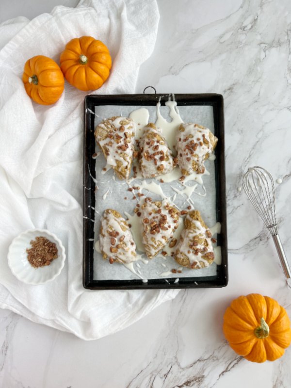 pumpkin scones with glaze and candied pecans, pumpkin scones on a black baking sheet with a whisk and pumpkins surrounding it