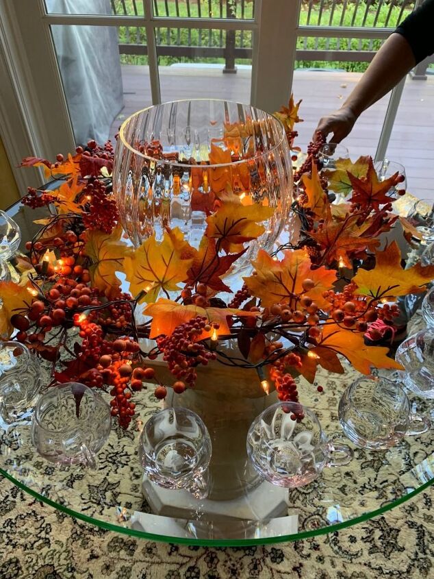 the best cider, A gorgeous Crate and Barrel punch bowl on a round glass table with clear glass cups Surrounding the punch bowl is a garland of silk leaves that are illuminated with white lights