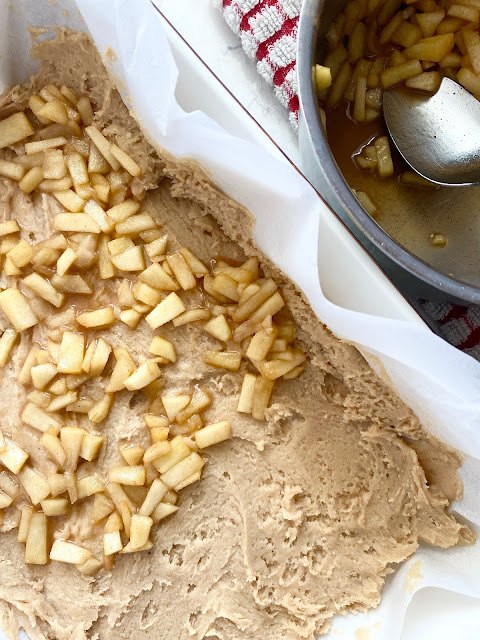 maple glazed apple blondies, Spiced apples laying over blondie batter with stainless steel bowl of apples and spoon