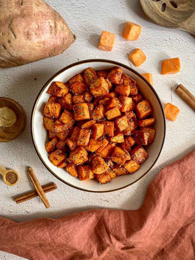 maple cinnamon air fryer sweet potato cubes happy honey kitchen, A bowl of air fried sweet potato chunks Whole and cubed sweet potatoes cinnamon sticks and measuring spoons with nutmeg and ginger surround the bowl