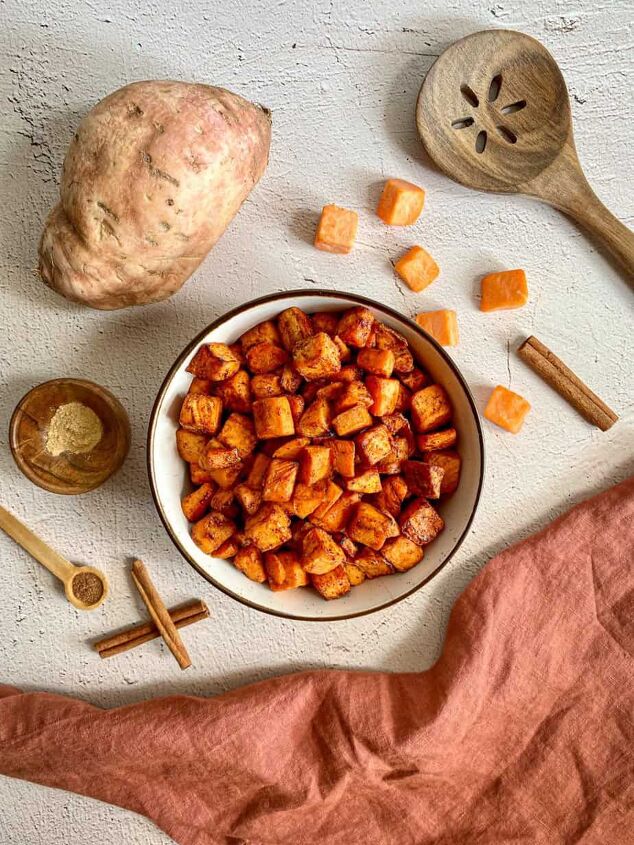 maple cinnamon air fryer sweet potato cubes happy honey kitchen, A bowl of air fried sweet potato chunks Whole and cubed sweet potatoes cinnamon sticks and measuring spoons with nutmeg and ginger surround the bowl A wooden spoon and rust colored towel are on the edges