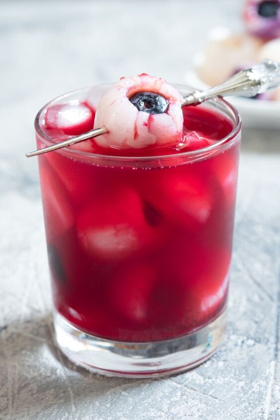blueberry lychee eyeball cocktail, Glass of Blueberry Lychee Eyeball Cocktail on a gray marble background with an eyeball garnish of a lychee stuffed with a blueberry to resemble a bloody eyeball