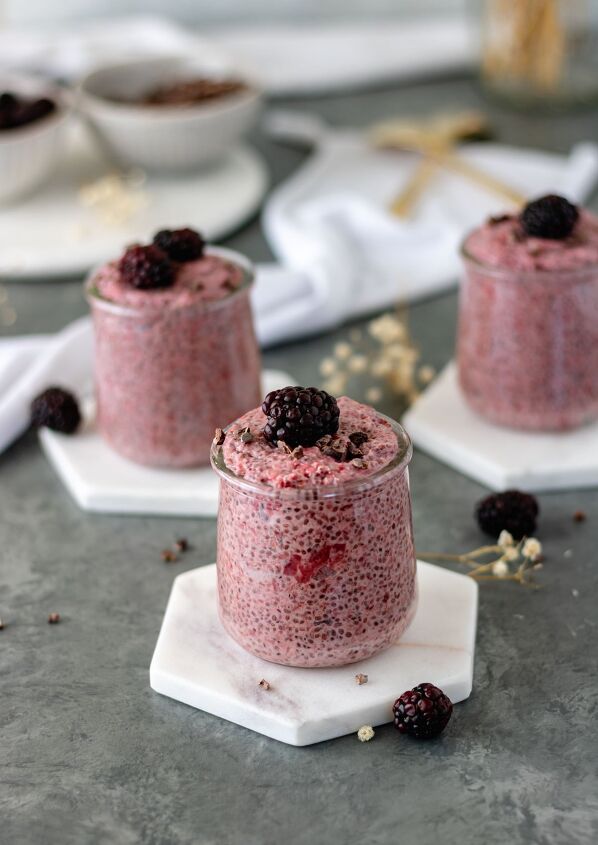 roasted strawberry chia pudding, Three mini glass jars of Roasted Strawberry Chia Pudding sitting on marble coasters alongside pinch bowls of cacao nibs and blackberries