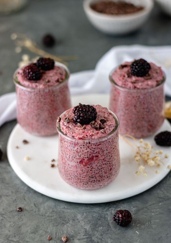 roasted strawberry chia pudding, Three mini glass jars of Roasted Strawberry Chia Pudding on a marble trivet and surrounded by cacao nibs blackberries and baby s breath