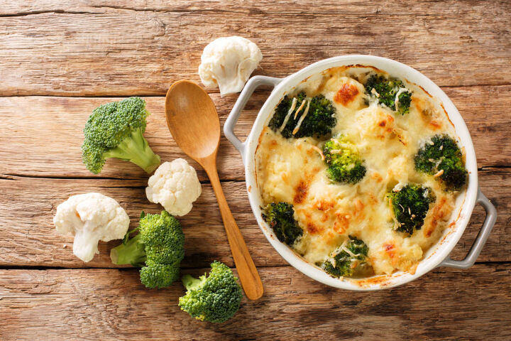 broccoli cauliflower alfredo casserole, wooden board with a round casserole of broccoli cauliflower alfredo casserole with raw broccoli and cauliflower florets on the side with a wooden spoon top view