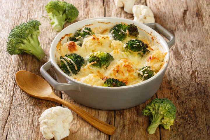broccoli cauliflower alfredo casserole, wooden board with a round casserole of broccoli cauliflower alfredo casserole with raw broccoli and cauliflower florets on the side with a wooden spoon