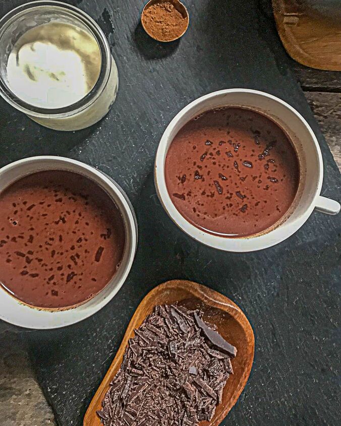 hot chocolate recipe without milk, two mugs of hot chocolate in a white mug a measuring spoon with ground cinnamon and pot of cream on a black slate board on a wooden table
