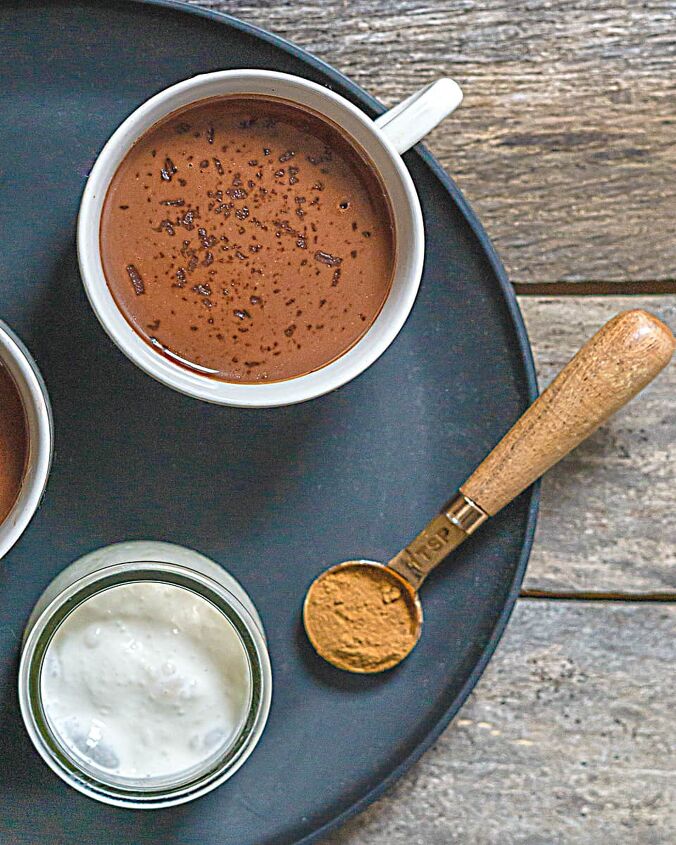 hot chocolate recipe without milk, dairy free hot chocolate in a white mug a measuring spoon with ground cinnamon and pot of cream on a black plate on a wooden table
