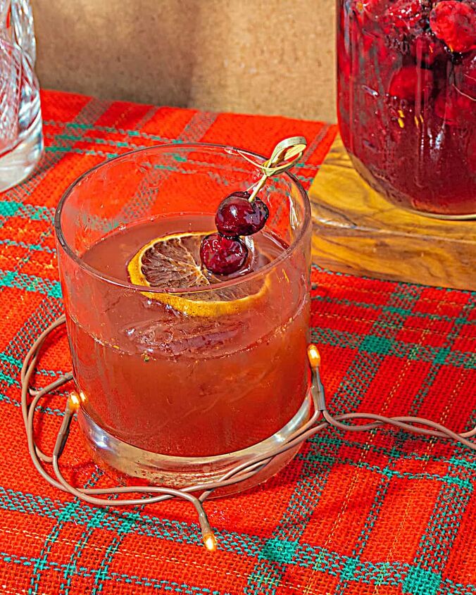 christmas old fashioned, old fashioned cocktail with a cocktail pick with dried orange slice and cranberries Jar of cranberry honey and mixing glass on background on a Christmas plaid tablecloth