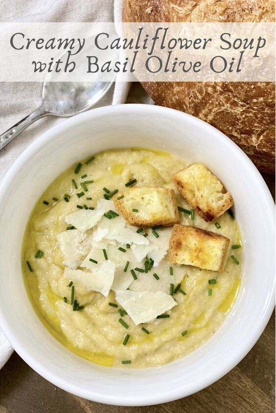 creamy cauliflower soup with basil olive oil, Creamy cauliflower soup with Basil Olive Oil in a white bowl garnished with parmesan cheese chives and croutons with a spoon and rustic bread on the side