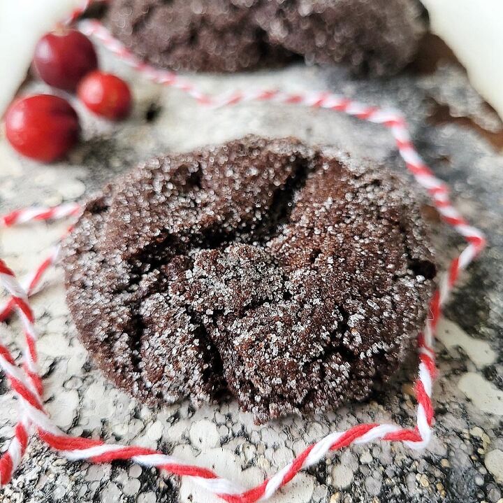 chocolate molasses crinkle cookies, functional image chocolate molasses cookies sprinkled with sugar front close up view of one cookie with two more in the background styled with cranberries and red and white holiday twine