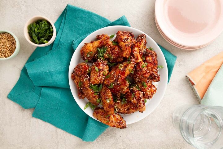 baked soy garlic chicken wings crispy, a large pile of saucy chicken wings on a white plate next to a stack of pink plates and a blue napkin