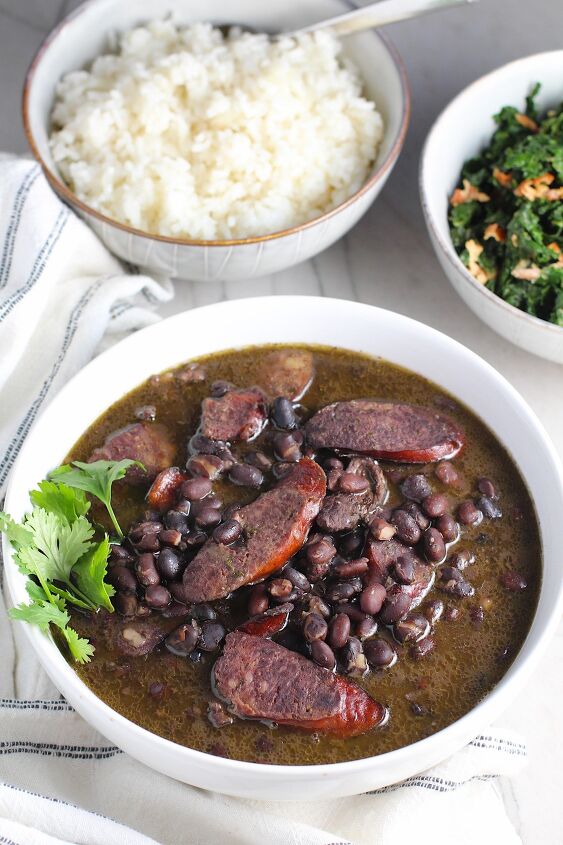 feijoada rice and beans brazilian style with meat, Bowl of cooked black beans with pork sausage slices and beef and cilantro garnish on side It is on table with a bowl of cooked kale and a bowl of rice with towel next to it Feijoada is rice and beans Brazilian style with pork sausages beef and more