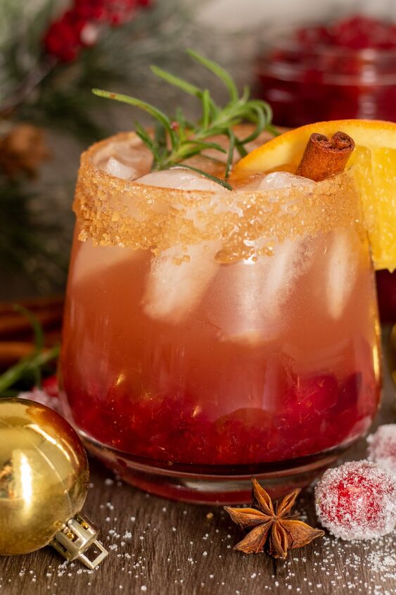 holiday cranberry bourbon cider, A short cocktail glass filled with holiday cranberry bourbon cider It s garnished with a sprig of rosemary orange slice and cinnamon stick It has a cinnamon sugar rim There s a gold ornament next to the glass with sugared cranberries and whole cinnamon sticks
