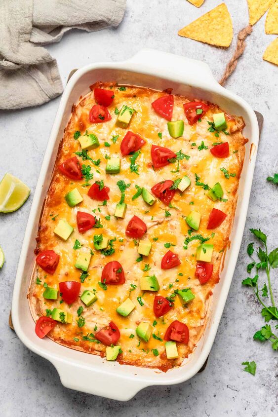 creamy chicken enchilada casserole, side angle shot of a taking a piece out of chicken enchilada casserole in a white baking dish with avocado and tomatoes