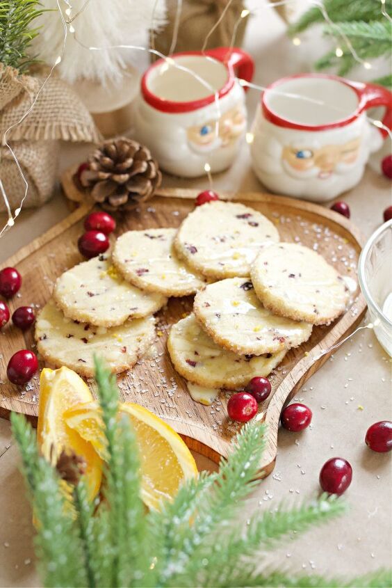 cranberry orange icebox cookies with orange zest drizzle, Cranberry orange cookies on a wooden ornament shaped cutting board with cranberries scattered around Santa cocoa cups and other Christmas decor