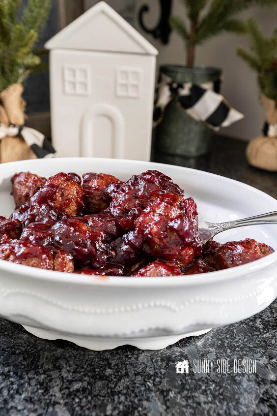Easy meatball appetizer in a cranberry sauce in a white serving dish with fork In the background are mini pine trees and a ceramic house