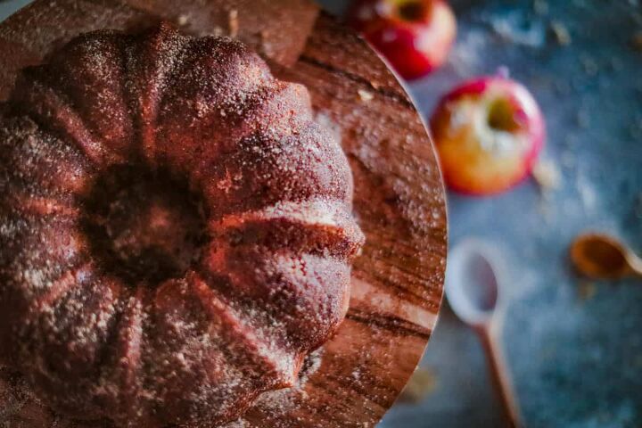 apple cider donut cake, overhead shot of this apple bundt cake on a cake stand with apples in the background