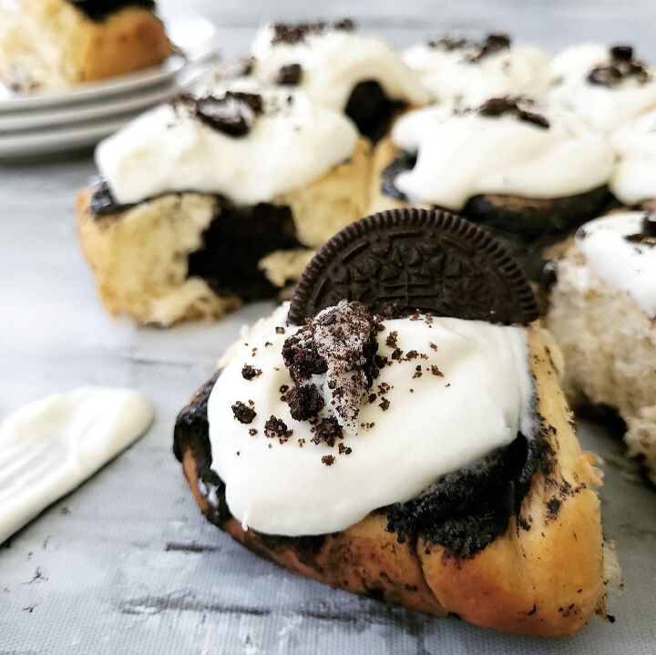 oreo cinnamon rolls, oreo cinnamon rolls full frame of cookies and cream cinnamon rolls one is in the background on a stack of plates all others are topped with cream cheese icing and oreo cookie crumbs one is featured in the front with half an oreo cookie propped on top