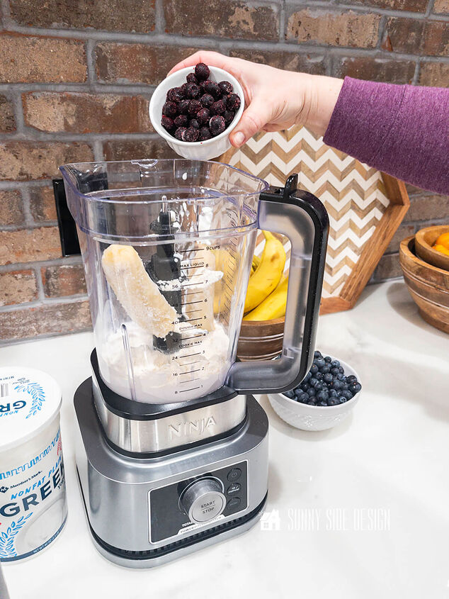 the best superfood blueberry banana smoothie, Woman s hand pouring a bowl of frozen blueberries into the blender for the healthy smoothie