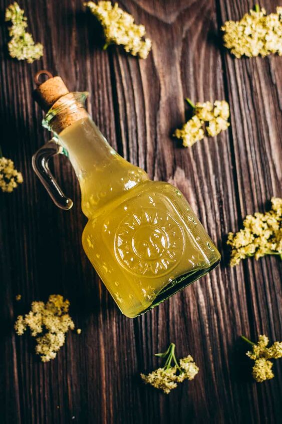 how to make elderflower cordial, fresh elderflower heads surround a glass sun bottle filled with golden syrup elder flower cordial recipe