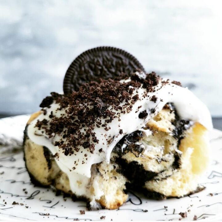 oreo brownies, oreo cinnamon roll side view of one roll with a bite missing to see the black cocoa swirled interior roll is topped with cream cheese and oreo crumbs background is abstract plate is white with i love you written in numerous languages