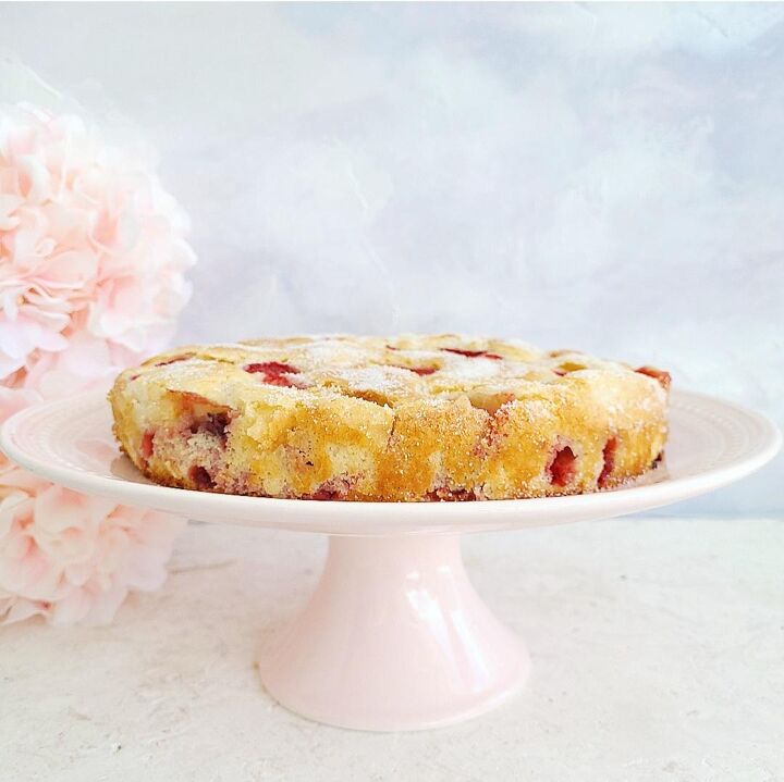 french strawberry cake, french strawberry cake uncut on a pale pink cake plate side view there are pink flowers stacked on the left side of the frame background is a wispy light blue and white abstract surface is white limestone