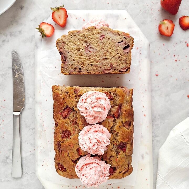 french strawberry cake, strawberry banana bread with strawberry buttercream rosettes top down view of loaf with one slice cut and facing upwards to showcase the crumb and strawberries loaf is on a rectangular piece of white marble on a gray slab and styled with freshly cut strawberries and a stainless steel knife