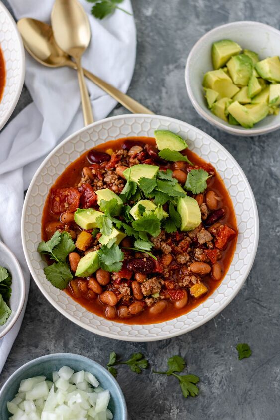 30 minute chicken chipotle chili, A glass ceramic bowl filled with Chicken Chipotle Chili and surrounded by pinch bowls of diced onion avocado and fresh cilantro