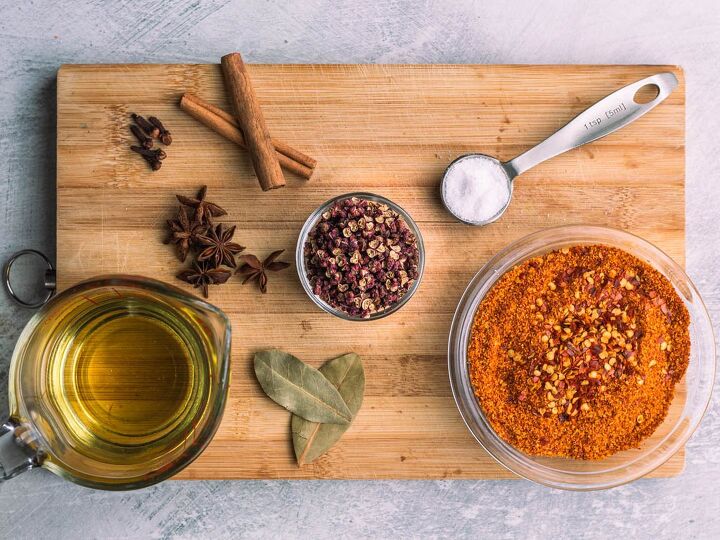 homemade spicy chili oil, cinnamon sticks a teaspoon measuring spoon with salt a small bowl with red powder two bay leaves a cup of oil five star anise five cloves on a brown cutting board on top of a light gray surface