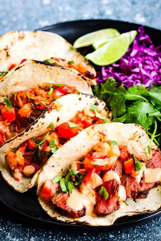 korean bulgogi tacos with kimchi salsa, vertical shot of 3 korean tacos corn tortillas filled with sliced steak kimchi and pico de gallo salsa and chopped cilantro In the background are lime wedges sliced red cabbage and cilantro leaves