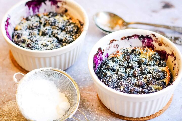 5 ingredient blueberry clafoutis for two, horizontal photo of blueberry clafoutis cooked in two small white ramekins They are sprinkled with powdered sugar and there are two spoons and a sieve with powdered sugar on the table next to the ramekins