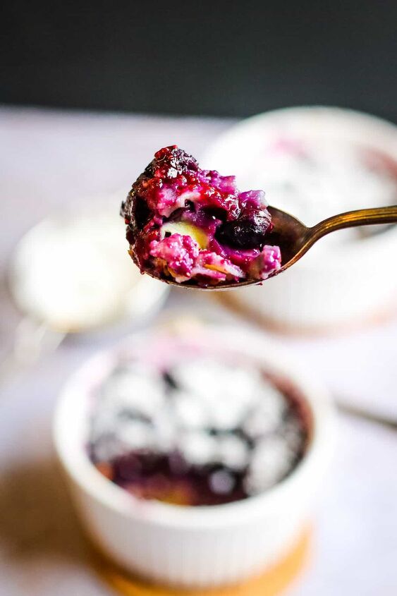 5 ingredient blueberry clafoutis for two, A low angle shot of blueberry clafoutis on a spoon In the background there is a blurred out ramekin of blueberry clafoutis topped with powdered sugar