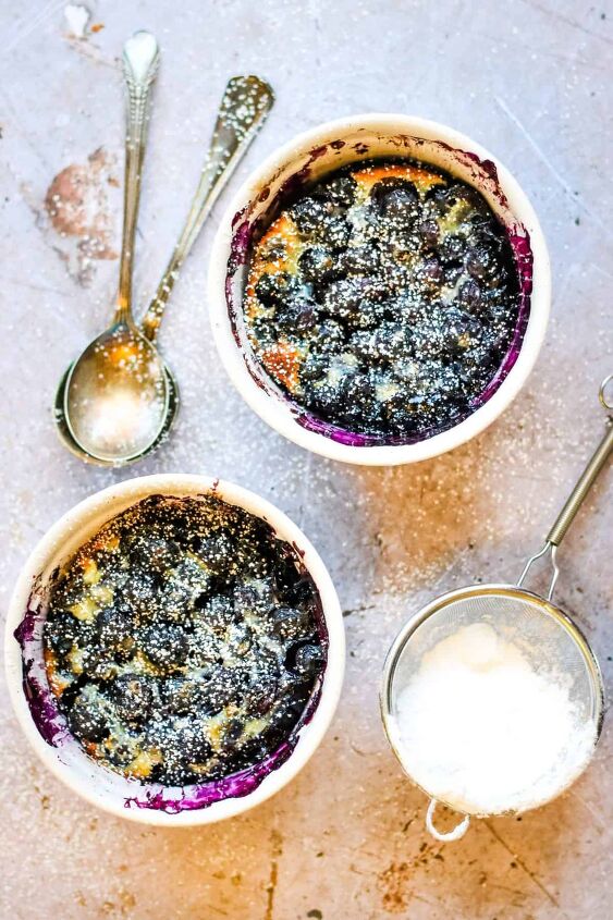 5 ingredient blueberry clafoutis for two, Overhead shot of two white ramekins with blueberry clafoutis topped with powdered sugar On the table next to the ramekins are two silver spoons stacked and a sieve with powdered sugar in it