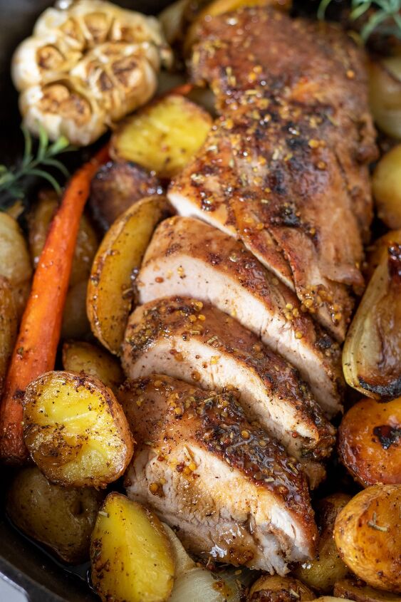 honey mustard roasted pork tenderloin with potatoes and carrots, A sliced pork tenderloin that s been baked in the oven It s got roasted vegetables in the cast iron skillet with it There s a head of garlic and fresh rosemary sprigs The pork tenderloin is brown on the outside and juicy on the inside