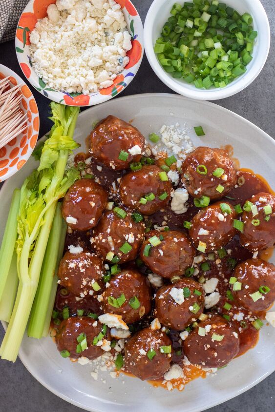 crockpot buffalo bbq chicken meatballs, A round white plate with bbq buffalo meatball appetizers There s topped with blue cheese crumbles and chopped scallions There s 3 small bowls in the background one with toothpicks one with scallions and one with blue cheese There s a couple of stalks of celery on the plate with the meatballs