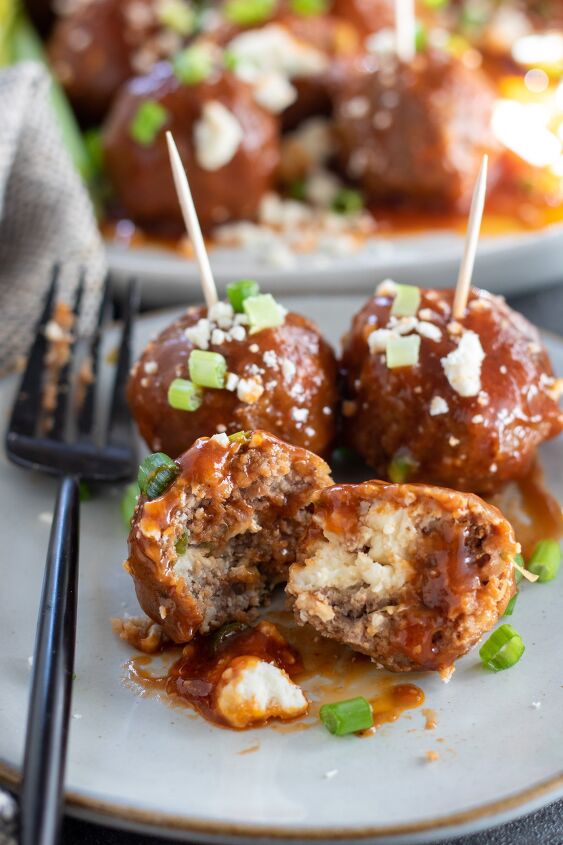 crockpot buffalo bbq chicken meatballs, A small plate with 3 small meatballs on it One meatball is cut open and it s stuffed with blue cheese The other two meatballs have toothpicks in them for appetizers There s a black fork on the plate and a bigger plate in the background with more buffalo chicken meatballs