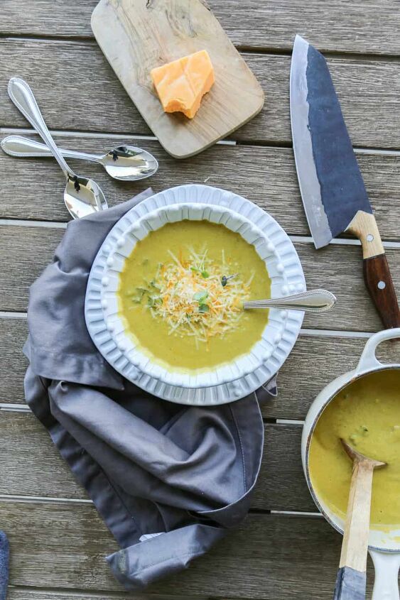gluten free broccoli cheddar soup keto soup recipe, gluten free broccoli cheddar soup in a bowl garnished with cheese and greens on a table with a napkin pot of soup and a knife and two spoons