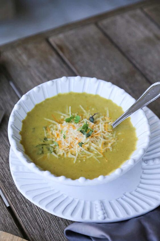 gluten free broccoli cheddar soup keto soup recipe, gluten free broccoli cheddar soup in a bowl garnished with cheese and greens on a table with a napkin pot of soup and a knife and two spoons