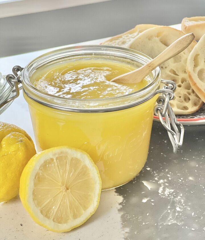 how to make lemon curd, A jar of lemon curd sitting on a counter beside cut lemons and a plate of english muffins