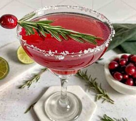 A cranberry margarita in a glass with a garnish of cranberries and a sprig of rosemary Fresh cranberries rosemary and limes surround the glass