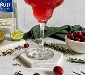 A Christmas margarita with a salt rim surrounded by fresh cranberries and limes A bottle of blanco tequila is behind the drink