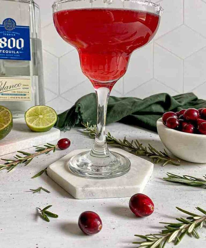 A Christmas margarita with a salt rim surrounded by fresh cranberries and limes A bottle of blanco tequila is behind the drink