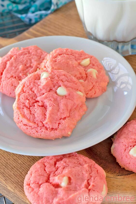 Strawberry Cake Mix Cookies are pink and dotted with white chocolate chips Cookies and a glass of milk are on a white plate that is resting on a wooden trivet