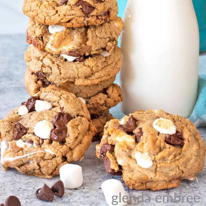 smores cookies stacked next to a milk bottle with a blue and white striped straw Two cookies are laying on the counter in front of the cookie stack and milk bottle with marshmallows and chocolate chips strewn around