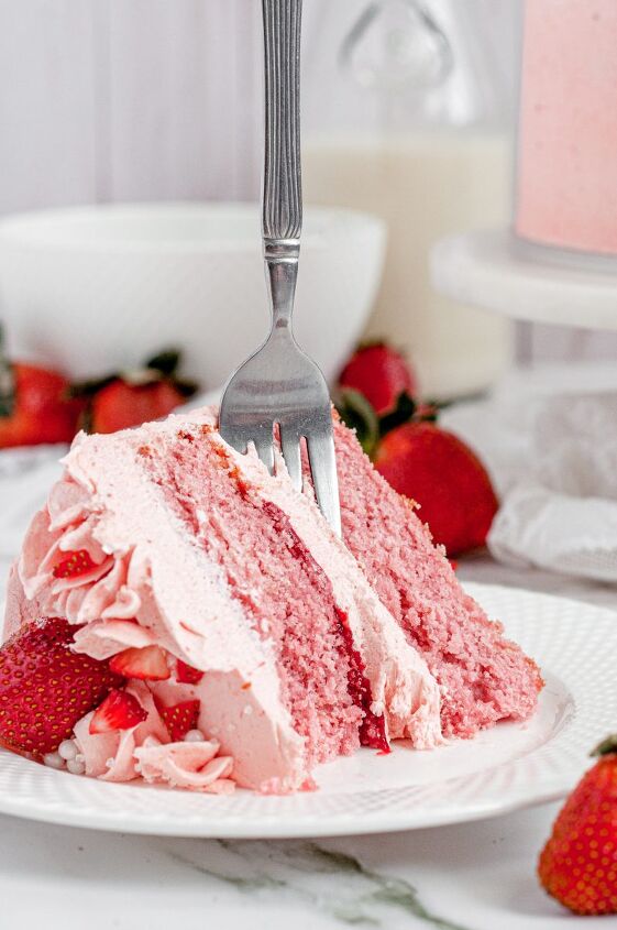 gluten free strawberry cake dairy free, A slice of pink strawberry cake sitting on a white plate It is topped with fresh red strawberries and covered in light pink frosting There is bright red strawberry filling in between the two cake layers and a fork is sticking in the cake slice straight up in the air