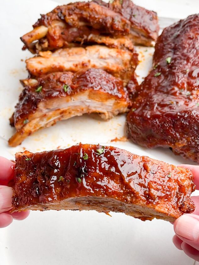 dr pepper crock pot ribs, hands holding one rib in foreground with racks of ribs in the background
