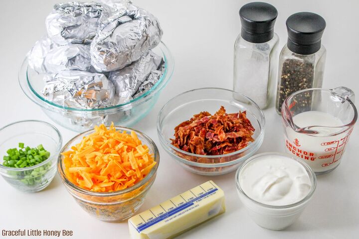 easy and delicious loaded baked potato casserole, A bowlful of foil wrapped baked potatoes a small bowl of shredded cheese a small bowl of crumbled bacon a small bowl of sour cream a stick of butter milk salt and pepper all sitting on a white counter